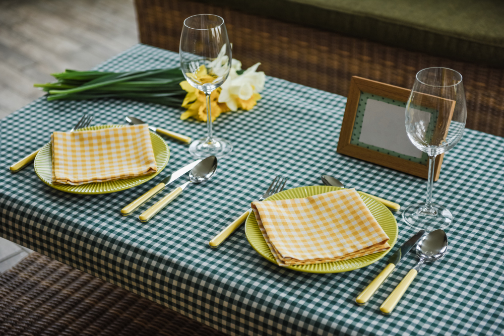 A table set for two with yellow checkered napkins, yellow-handled cutlery, empty wine glasses, daffodils, and a blank picture frame on a green checkered tablecloth.