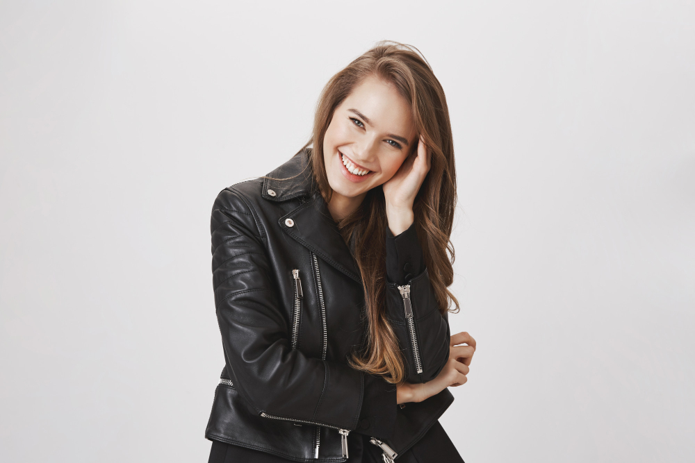 A young woman with long hair wearing a black leather jacket smiles and touches her hair while standing against a plain light background.