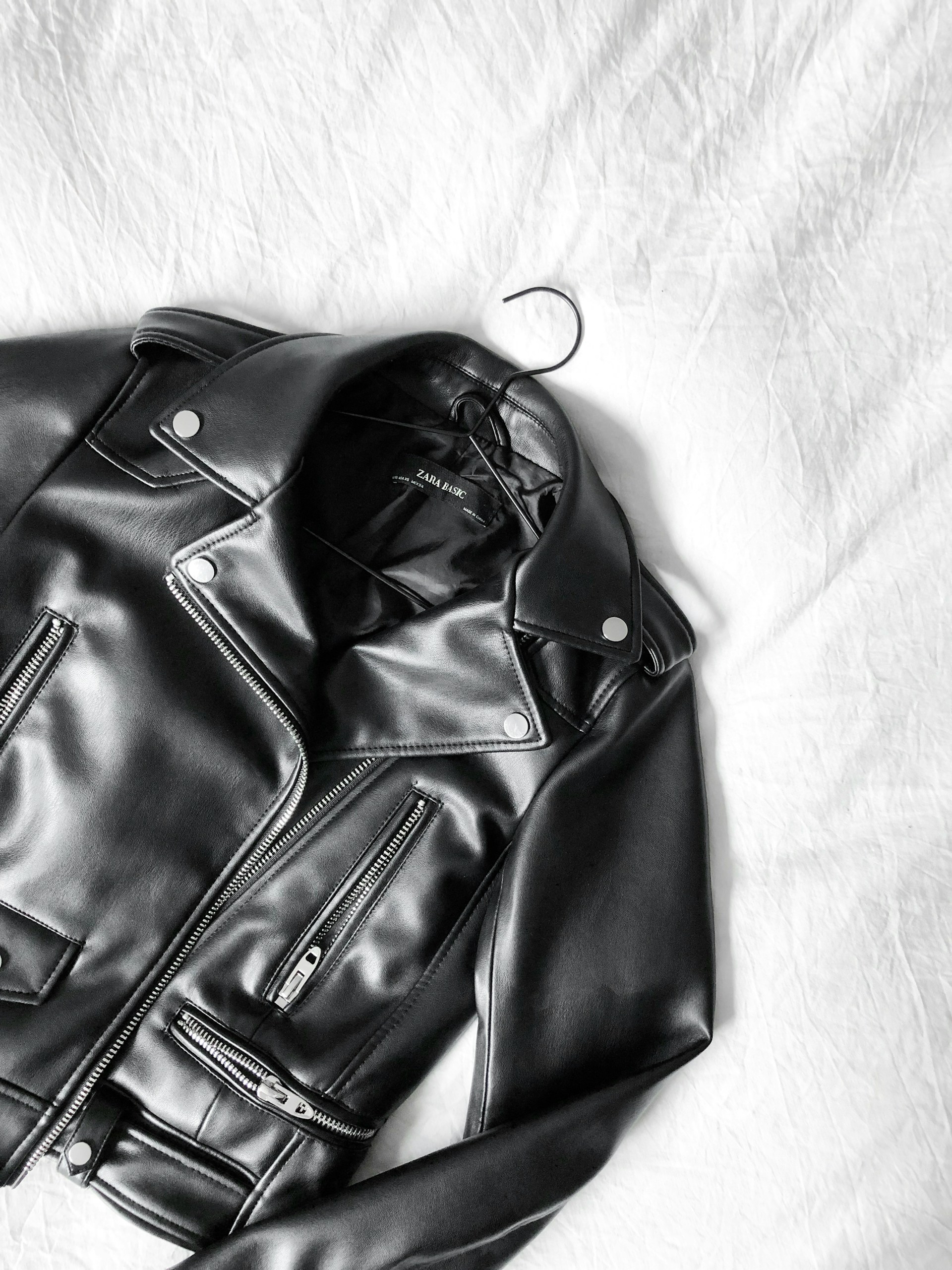 A black faux leather biker jacket with silver zippers and buttons is laid flat on a white sheet with a hanger visible at the collar.