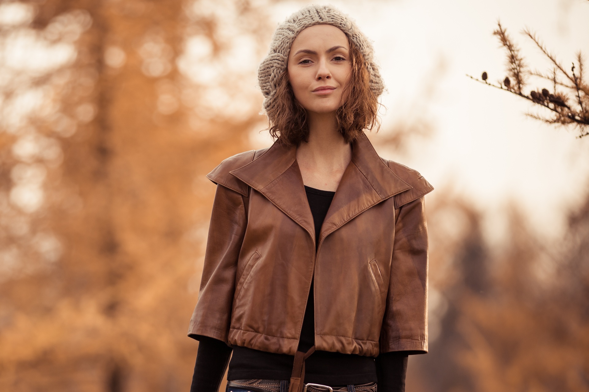 A woman wearing a knit hat and brown leather jacket stands outdoors with autumn-colored trees in the background.