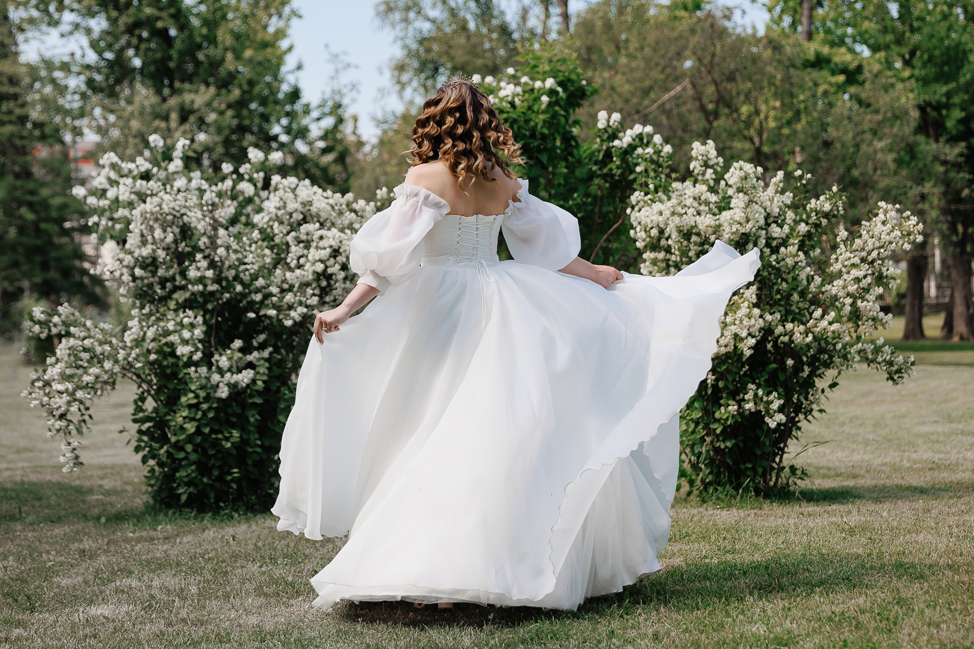 A woman in a white, off-the-shoulder, puff-sleeved gown twirls on grass in front of blooming white bushes on a sunny day.