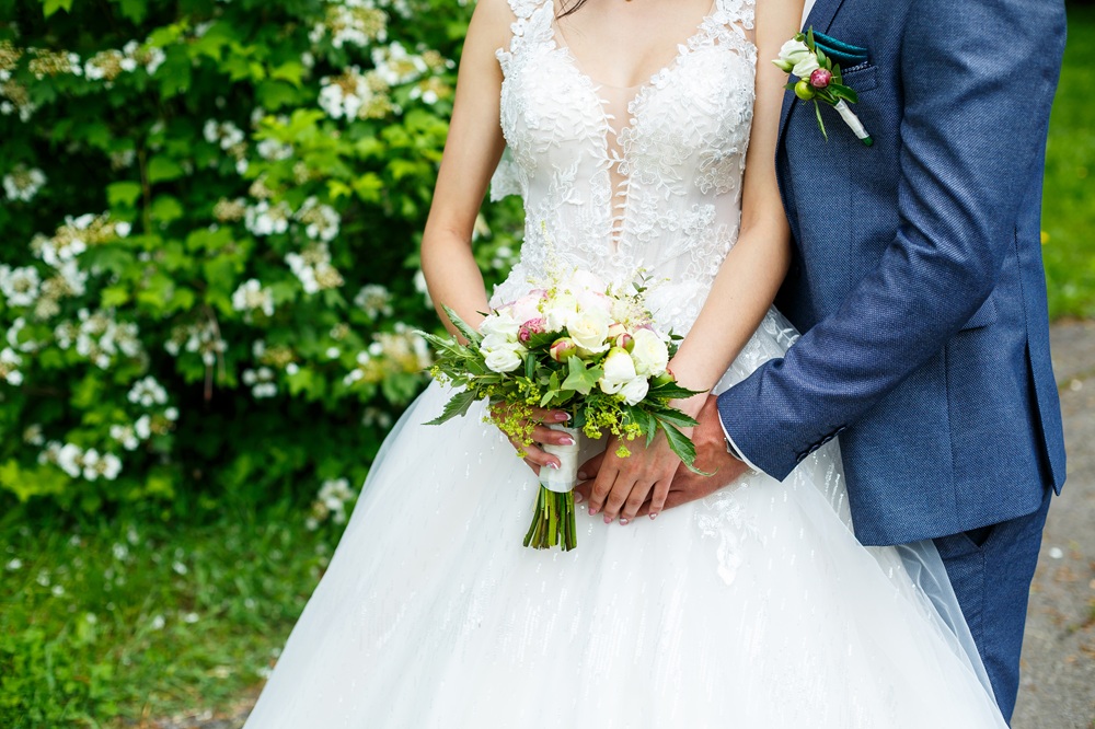 Bride in a white lace wedding dress holds a bouquet of flowers, standing next to a groom in a blue suit with a boutonnière, outdoors near greenery.