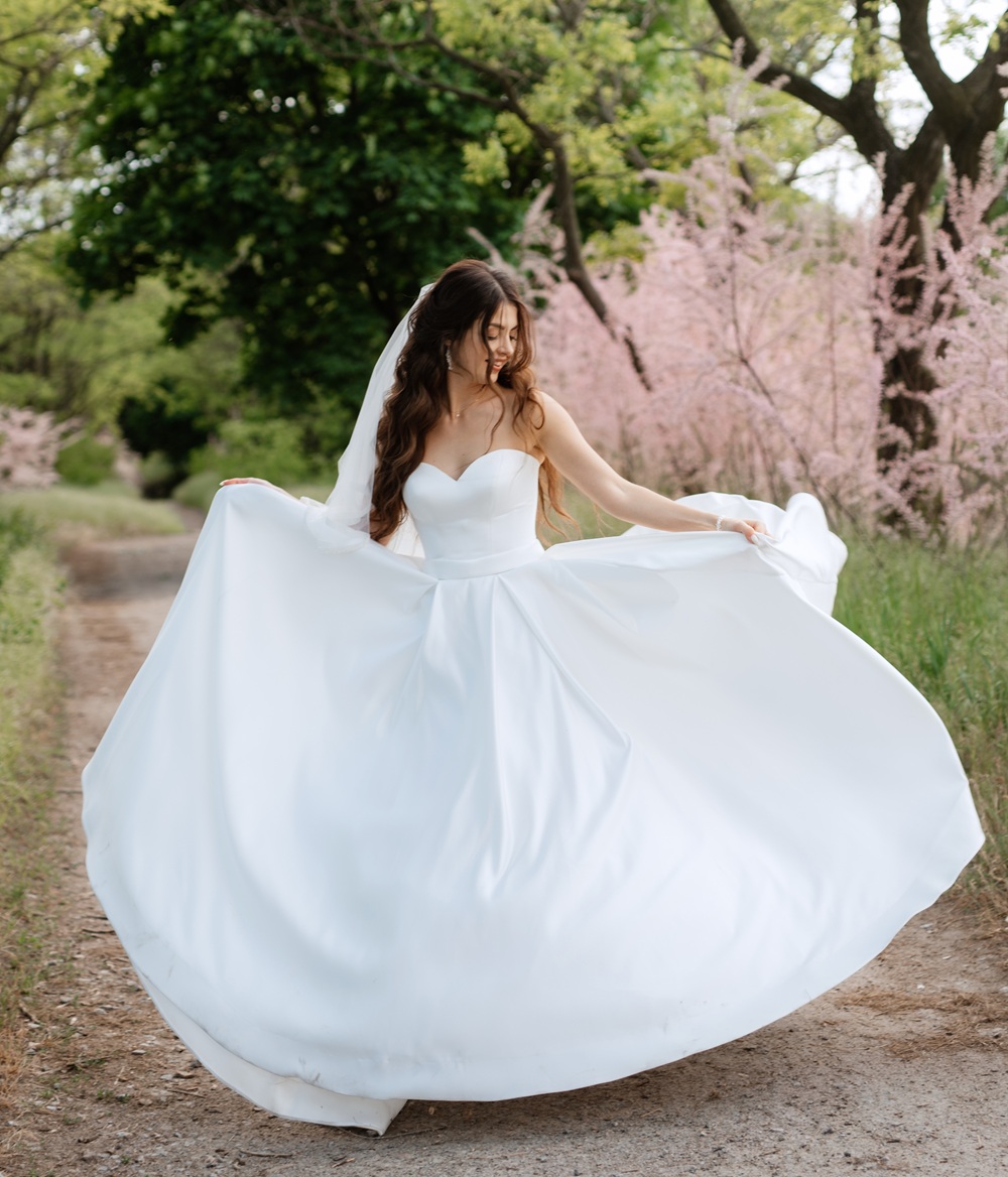 A woman in a strapless white wedding dress and veil twirls on a tree-lined path with blooming pink flowers in the background.