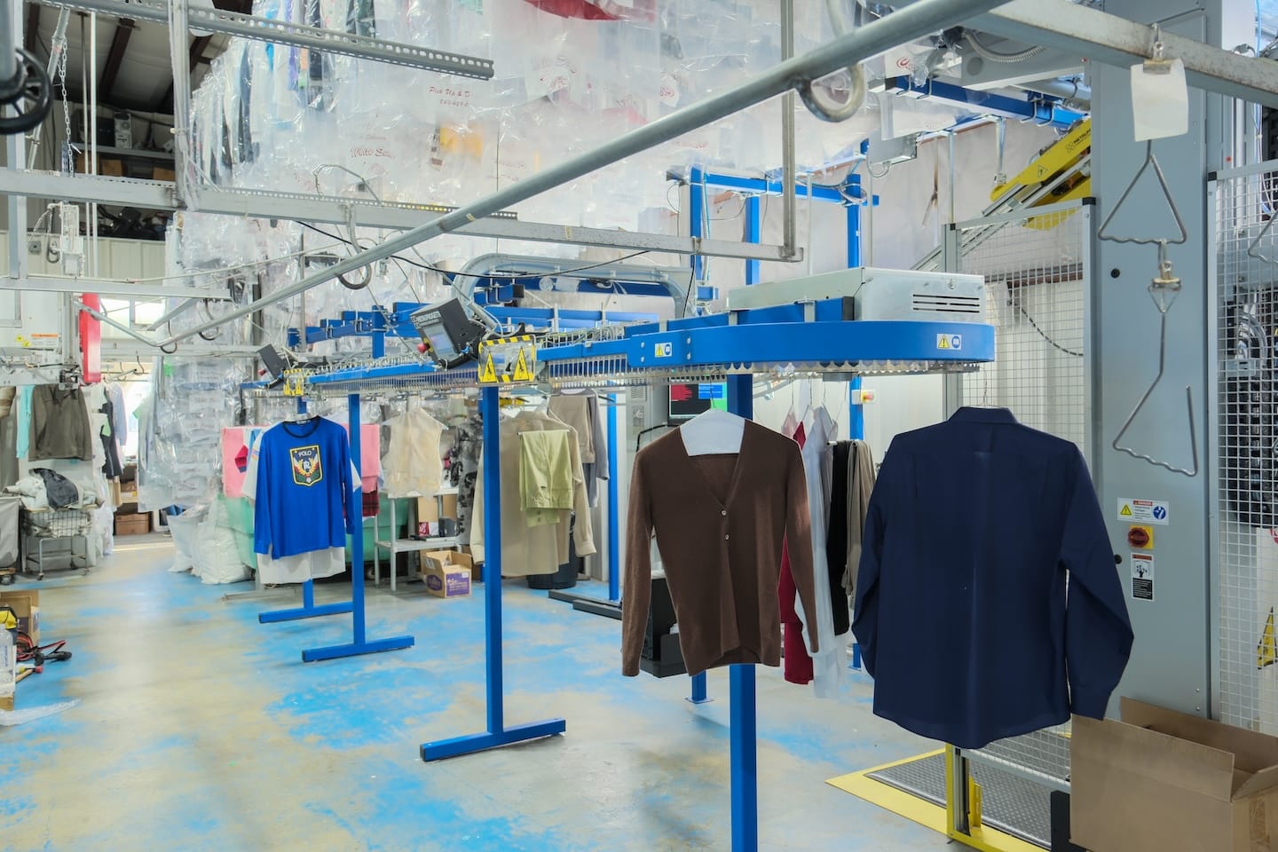 Clothes are hanging on a conveyor system in a dry cleaning facility, with plastic covers on garments in the background and various equipment visible.