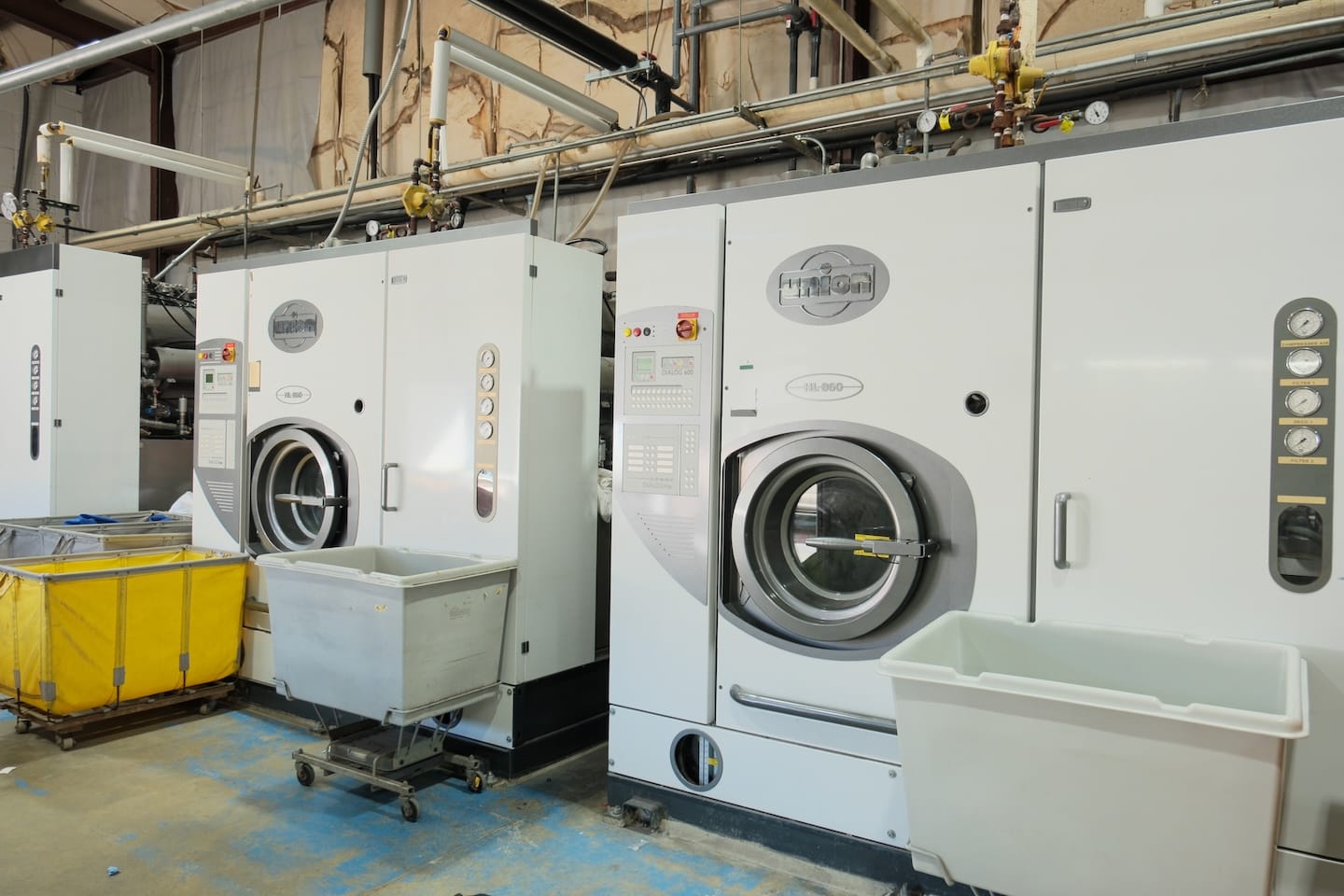 Two large industrial washing machines are positioned side by side in a laundry facility, with plastic carts and bins in front of them on a blue and beige floor.