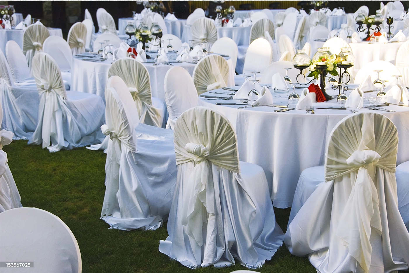 Round tables with white tablecloths and chairs covered in white fabric are set outdoors on grass, arranged for a formal event or celebration.