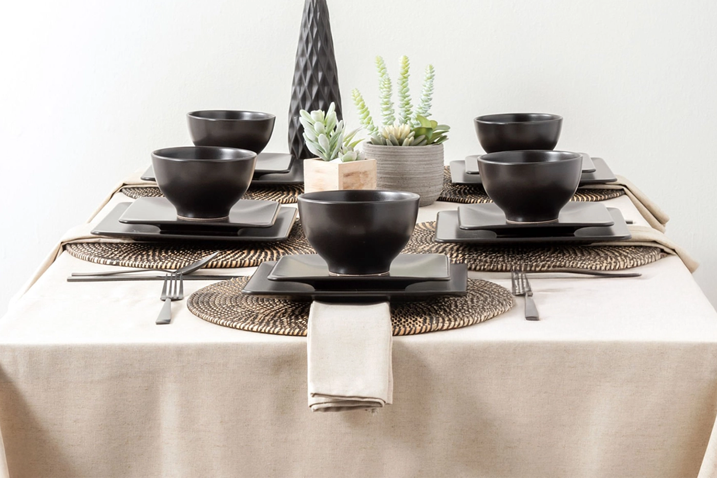 A dining table set for six with black dishware, woven placemats, beige napkins, and small potted succulents as a centerpiece.