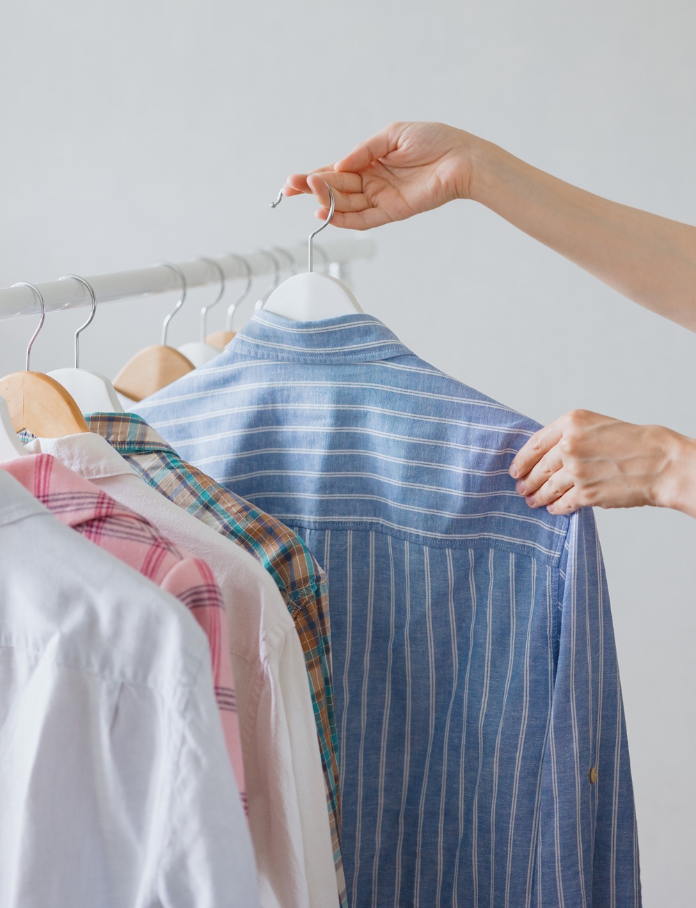 A hand selects a blue striped shirt from a clothing rack with several shirts, including plaid and solid patterns, on hangers.