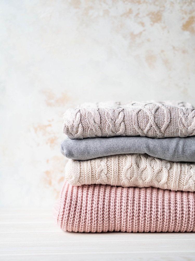A neatly folded stack of four sweaters in neutral and pastel colors, placed on a light wooden surface with a textured beige background.