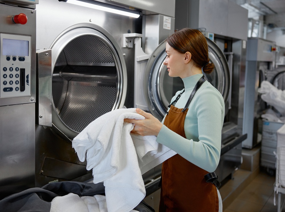A woman wearing an apron loads white towels into an industrial washing machine in a commercial laundry facility.