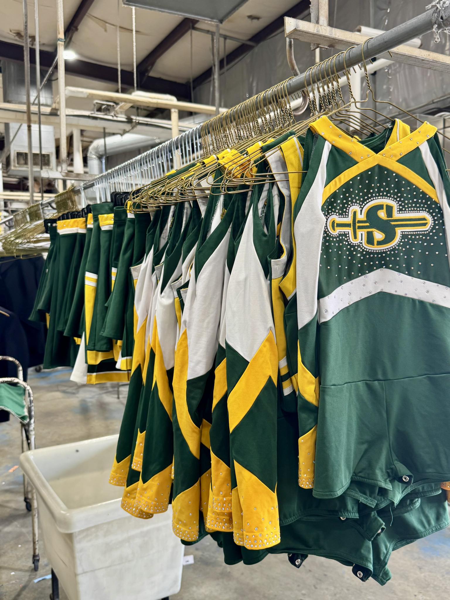 Green, yellow, and white cheerleading uniforms with an "S" logo hang on a rack in what appears to be a laundry or storage facility.