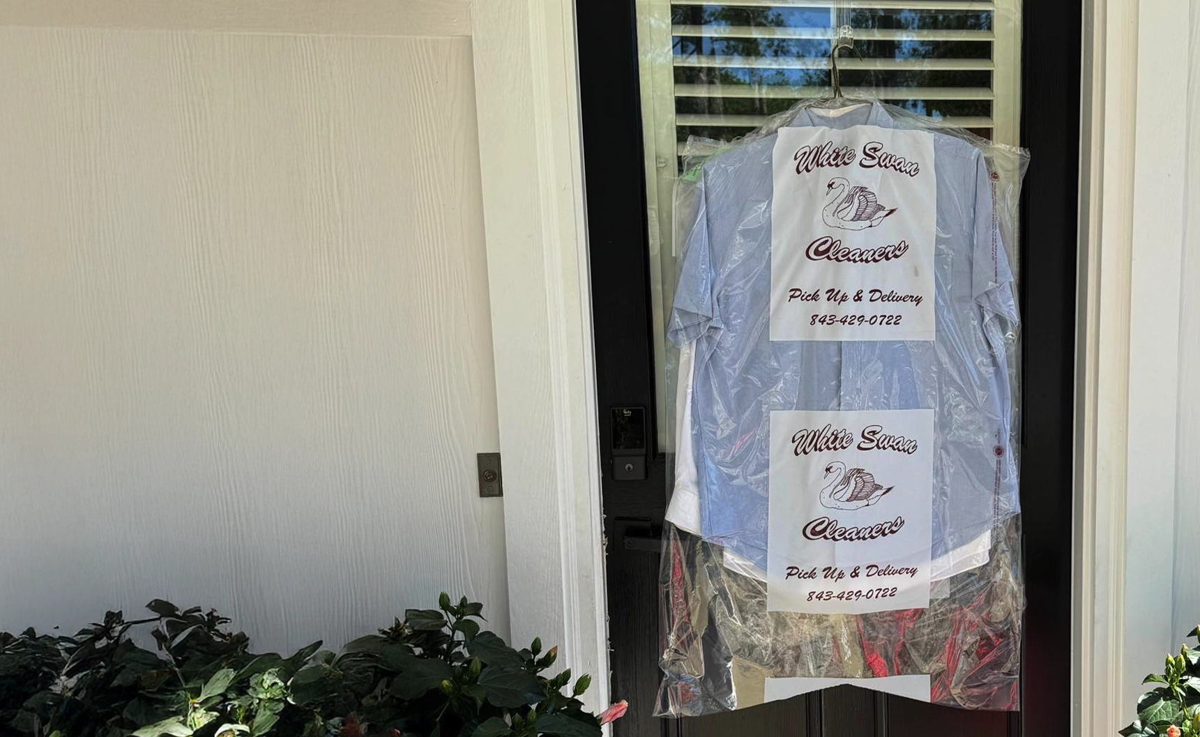 A plastic-wrapped shirt from a dry cleaner labeled "Mike Swan Cleaners" hangs on a front door against a white wall with plants in the foreground.