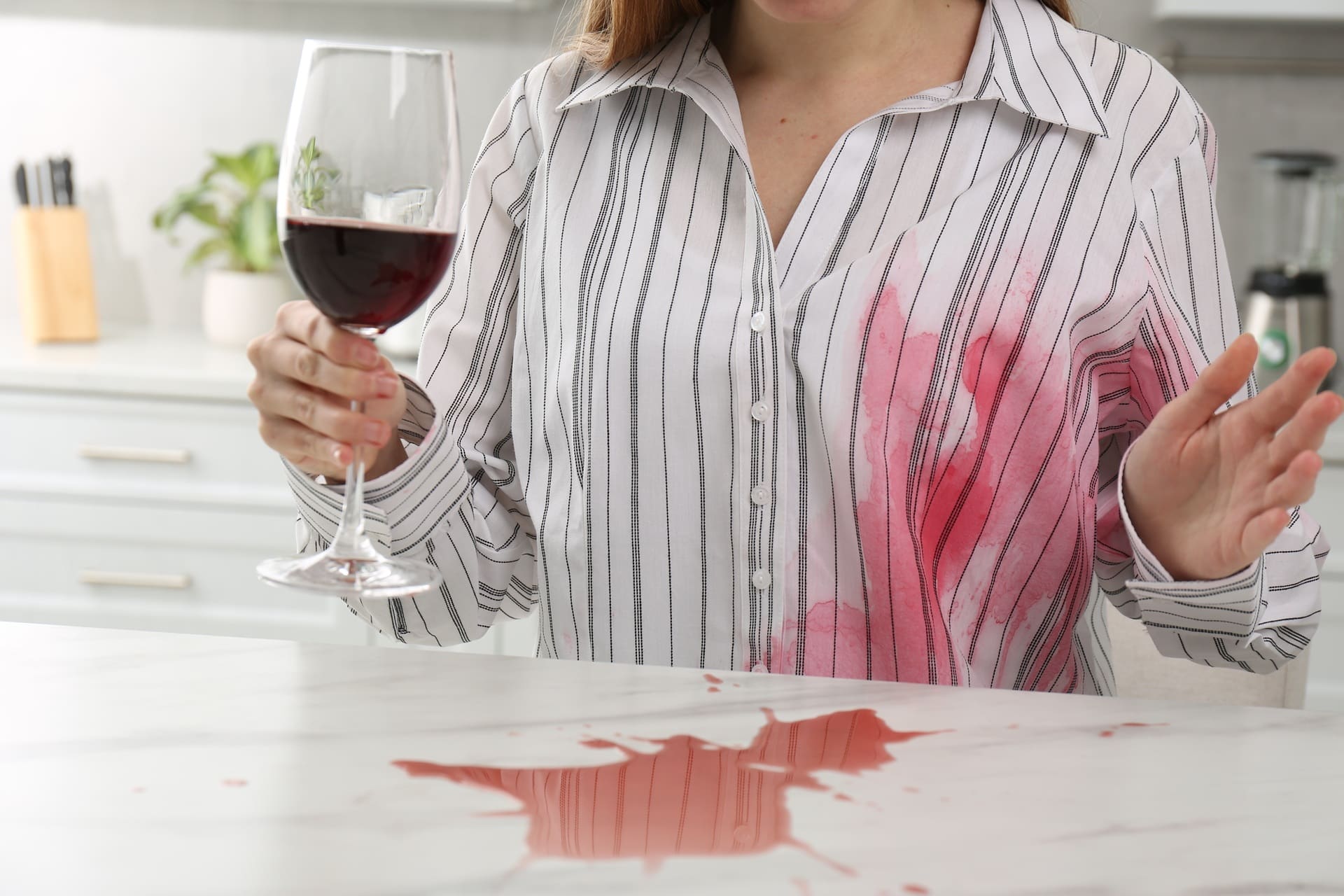 A person holding a wine glass has spilled red wine on their striped shirt and a kitchen counter.