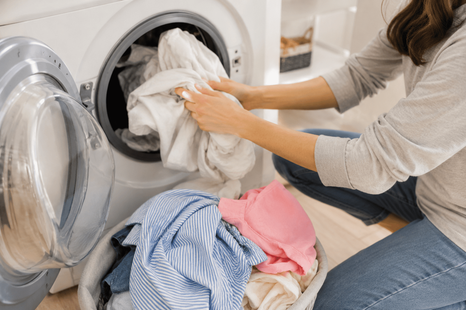 Person loading white laundry into a front-loading washing machine, with a basket of assorted clothes on the floor nearby.