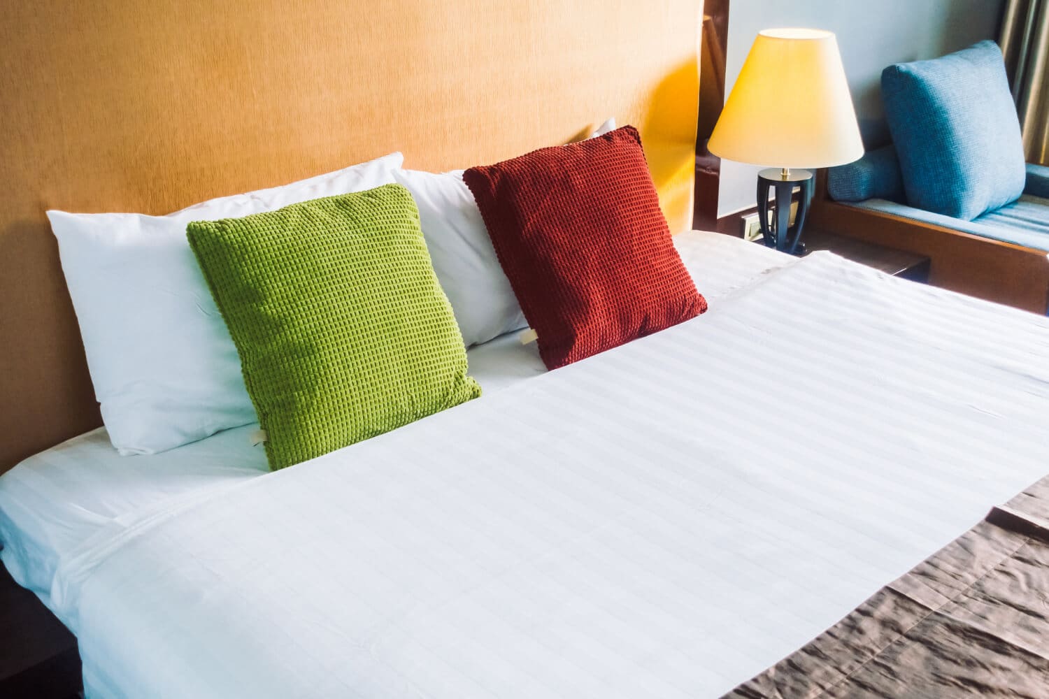 A bed with white linens, one lime green square pillow, and one red square pillow, with a lamp on a side table nearby.
